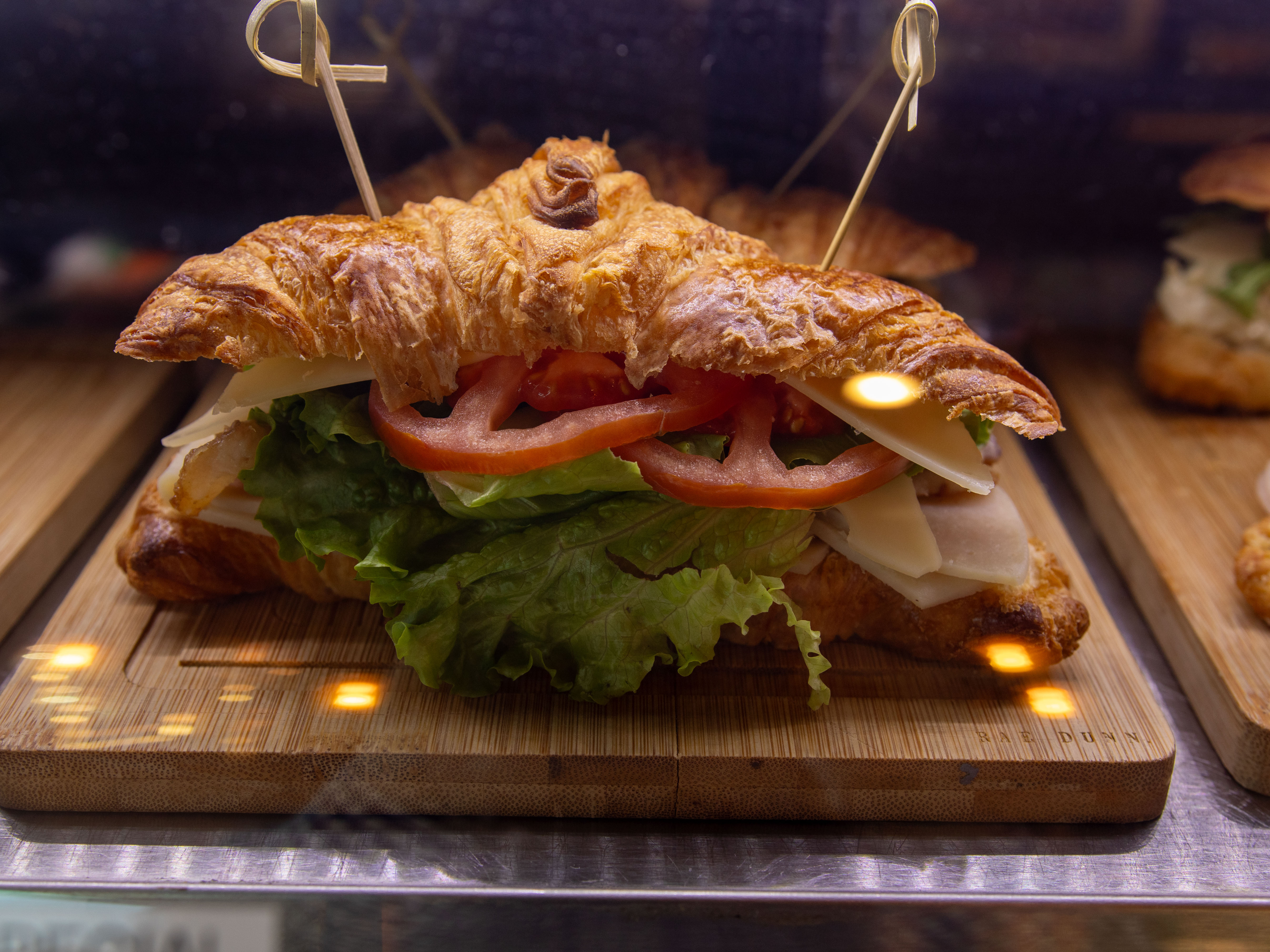 A close-up of a croissant sandwich filled with lettuce, tomato, and slices of cheese and meat, displayed on a wooden board.