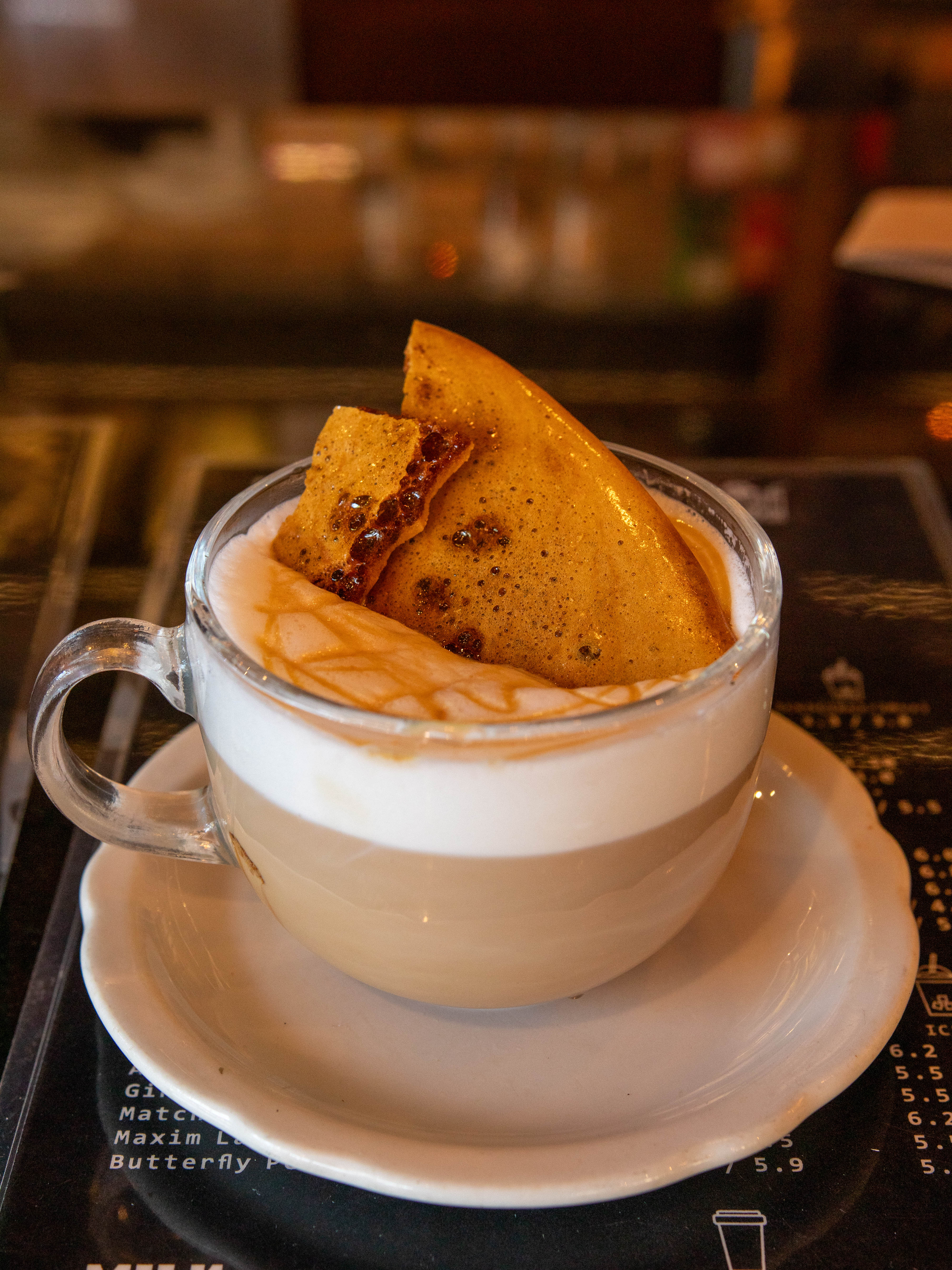 A close-up of a layered drink in a glass cup topped with foam and a decorative piece, placed on a white saucer.
