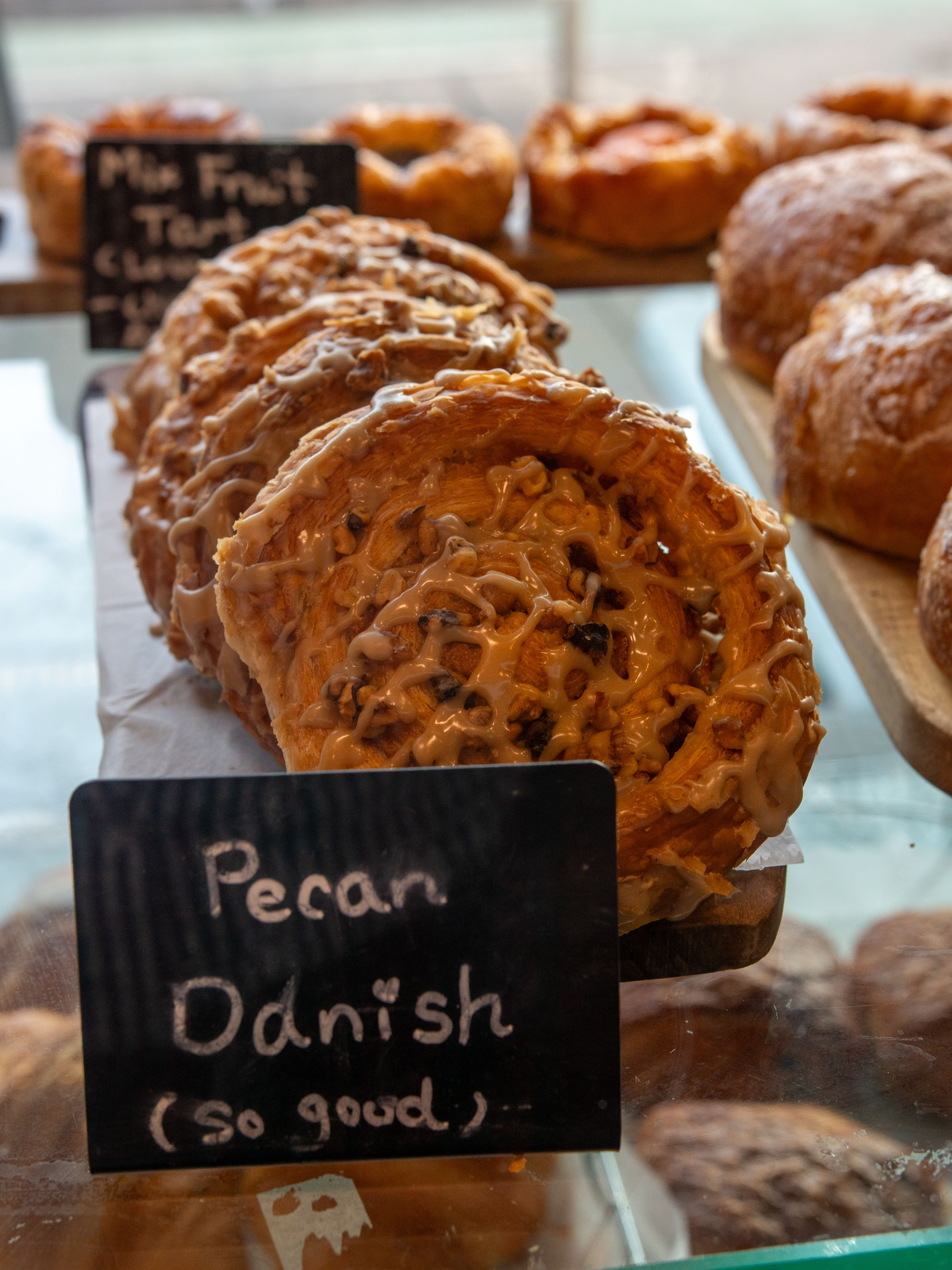 A display of freshly baked Pecan Danish pastries, topped with caramel icing, alongside a sign that reads 'Pecan Danish (so good)'